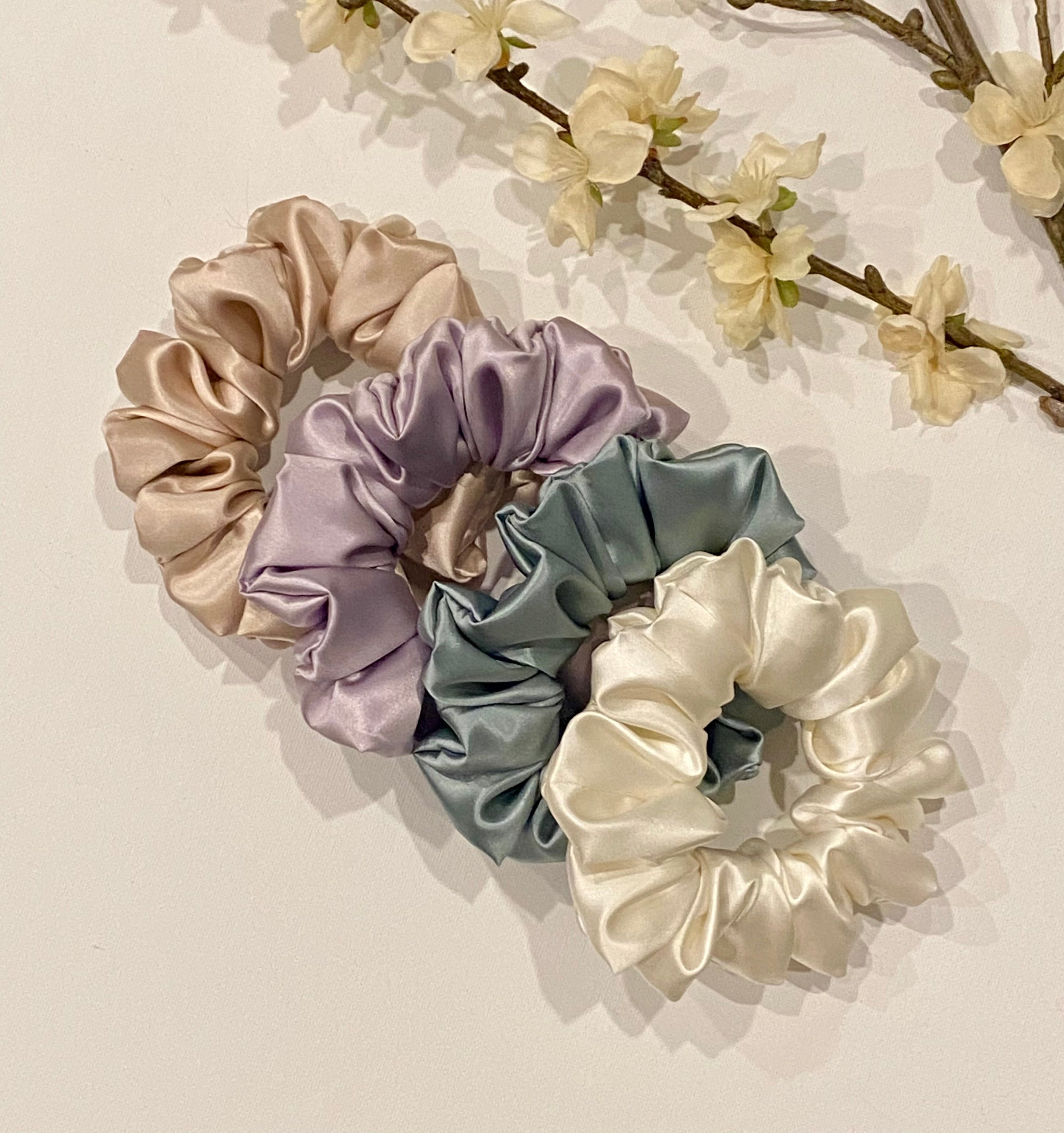 Set of four satin scrunchies in beige, purple, blue, and white on a light background with floral elements.